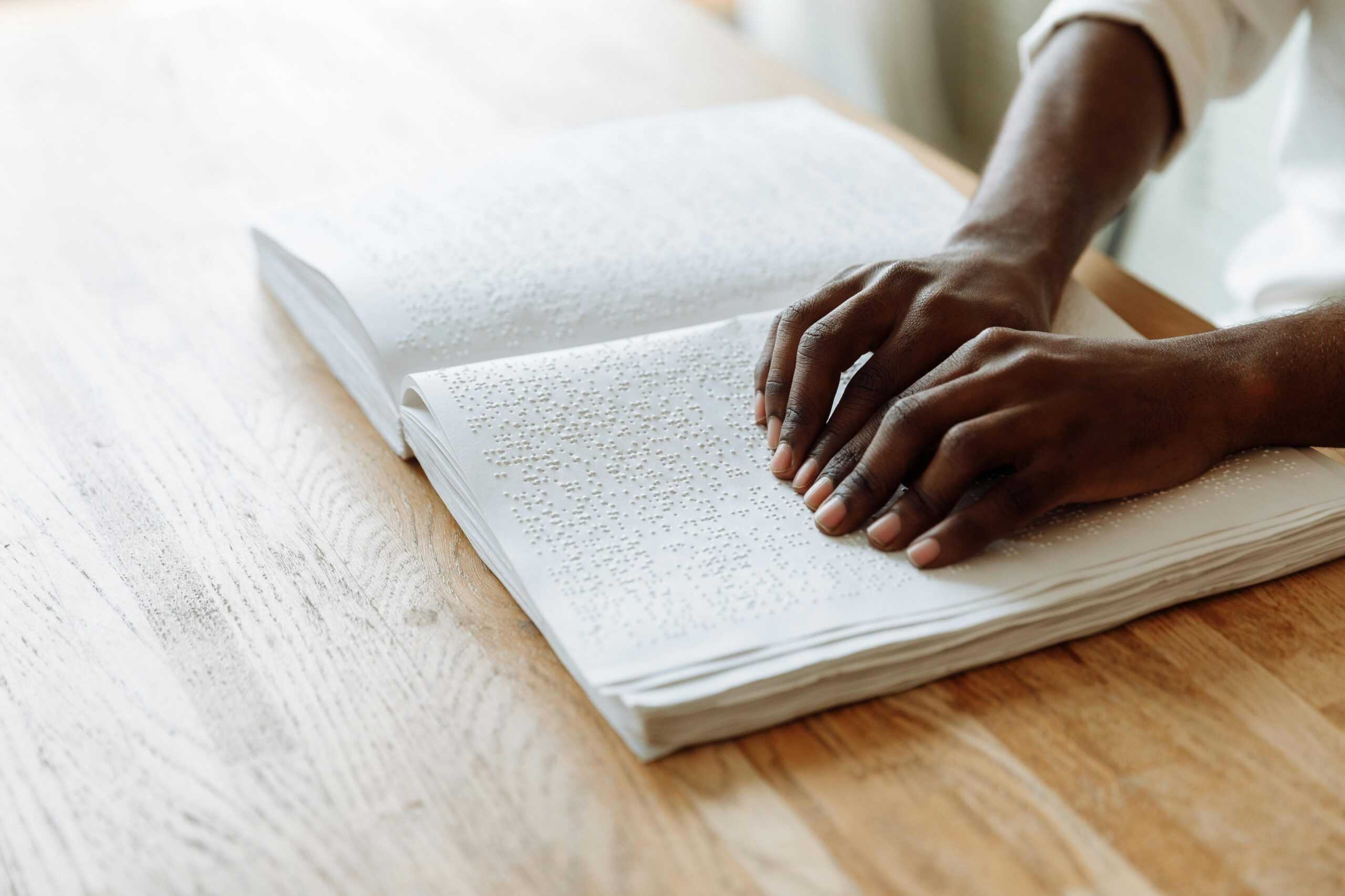 pexels photo 7265361 7265361 scaled Hands exploring a Braille book on a wooden table, emphasizing accessible learning.